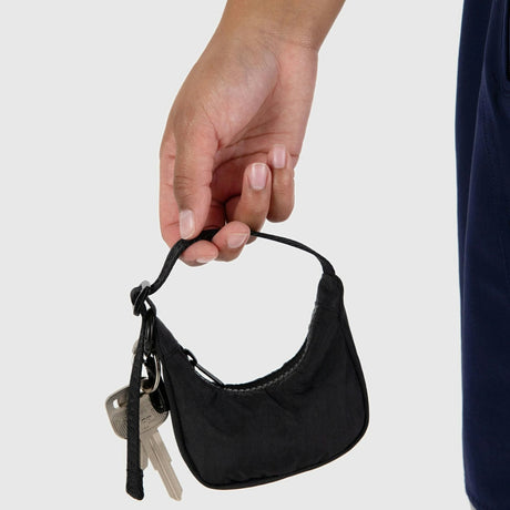 A hand holds the sleek black Baggu Crescent Bag crafted from recycled nylon, elegantly adorned with a set of keys and a Baggu Cornflower Charm.