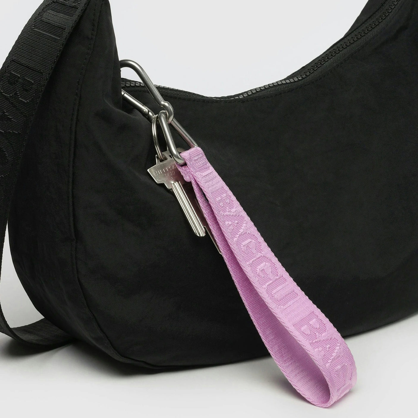 Black handbag with a Baggu Logo Keychain - Peony attached, featuring a silver key and a purple strap that reads "BAGGU." The keychain is crafted from recycled nylon webbing, ensuring durability and sustainability.