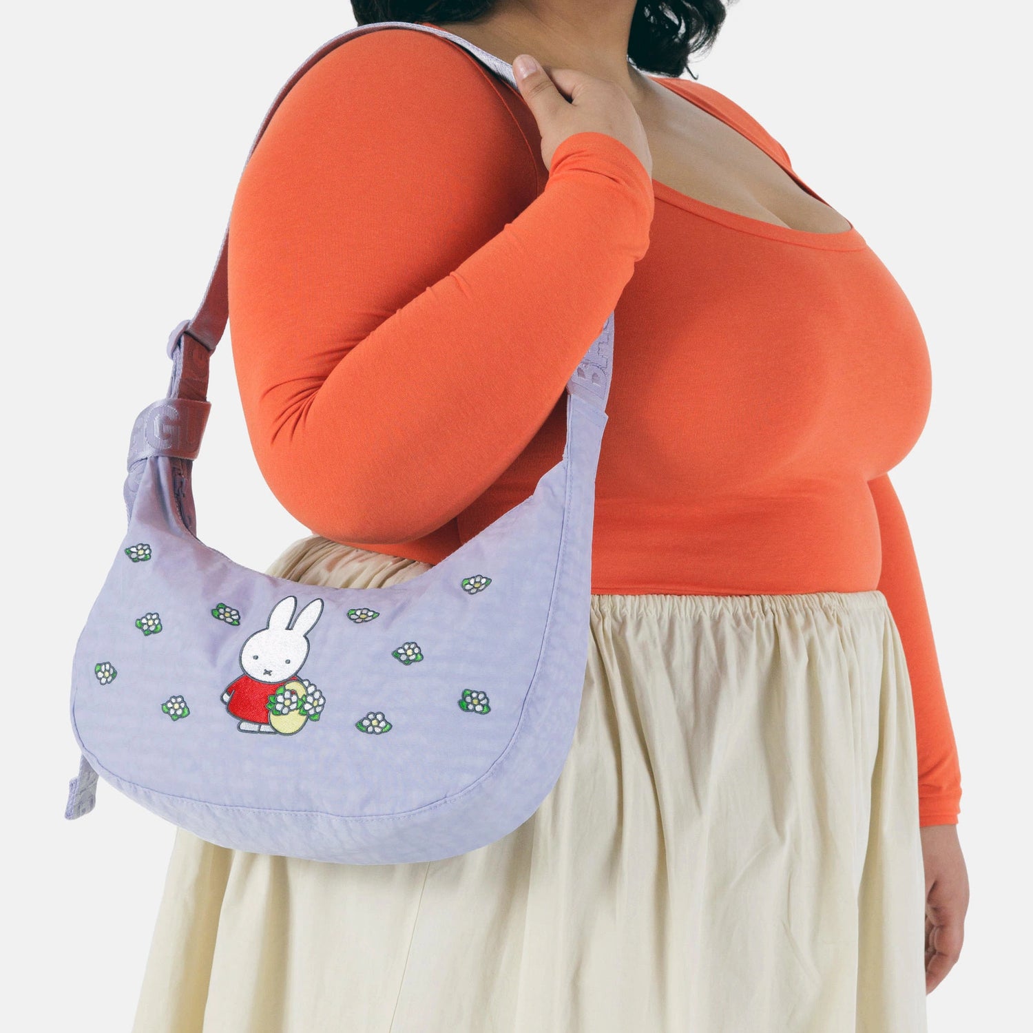A person in an orange top and cream skirt holds the Baggu Miffy x Baggu Medium Nylon Crescent, featuring a cartoon bunny and flowers.
