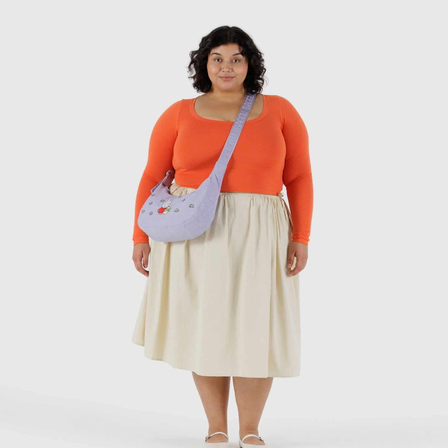 A woman faces forward, wearing an orange long-sleeve top, cream skirt, white shoes, and the Baggu Miffy x Baggu Medium Nylon Crescent crossbody bag in lavender with small embroidered details.