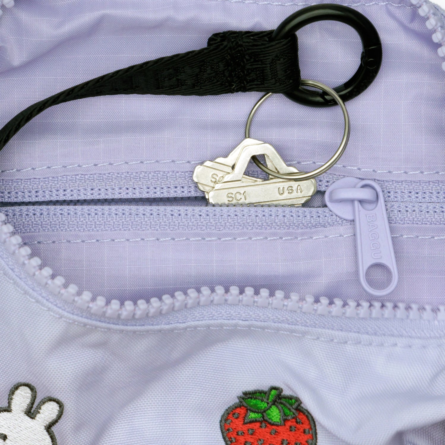 Close-up of two keys on a keyring inside the Baggu Miffy x Baggu Small Nylon Crescent bag in light purple, featuring embroidered patches such as a strawberry and Miffy.
