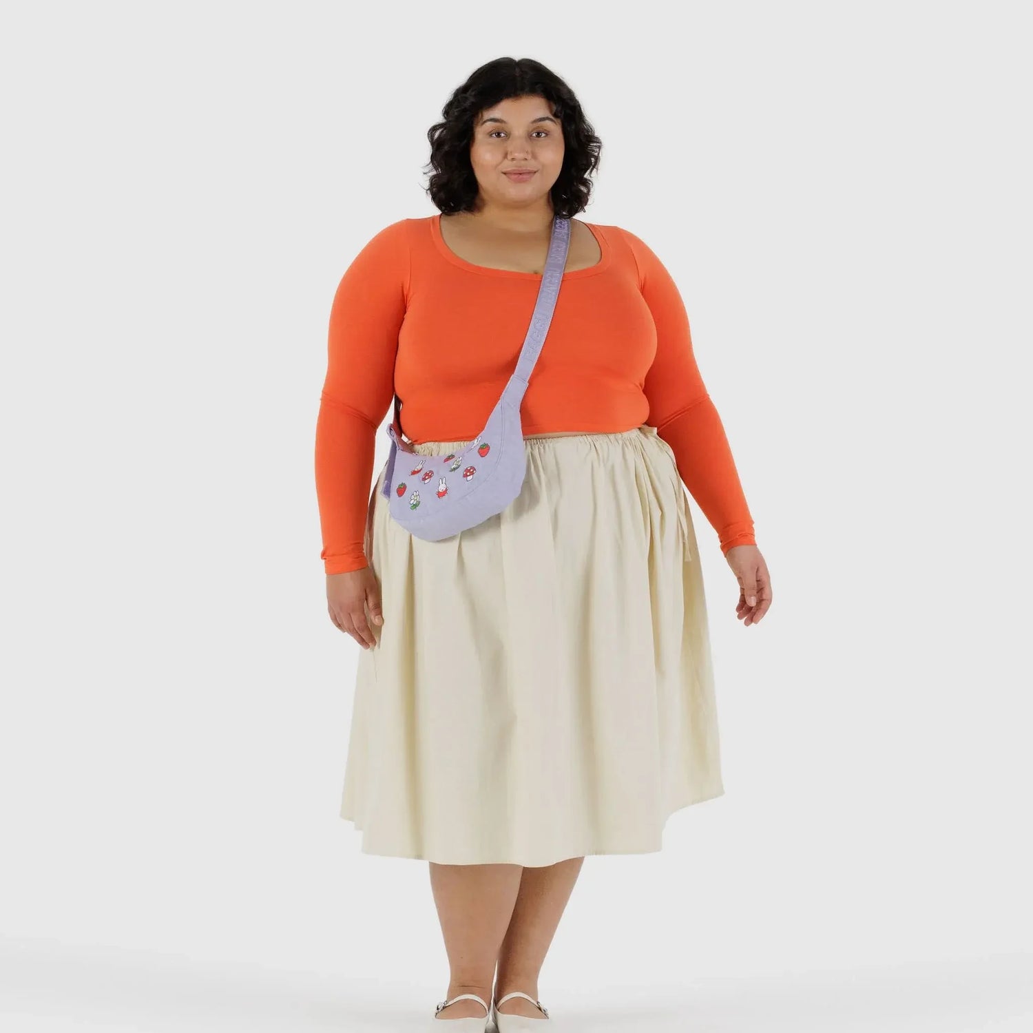 A woman faces forward, wearing an orange long-sleeve top, cream skirt, white shoes, and carries the Baggu Miffy x Baggu Small Nylon Crescent bag in light purple, featuring charming Miffy embroidery.