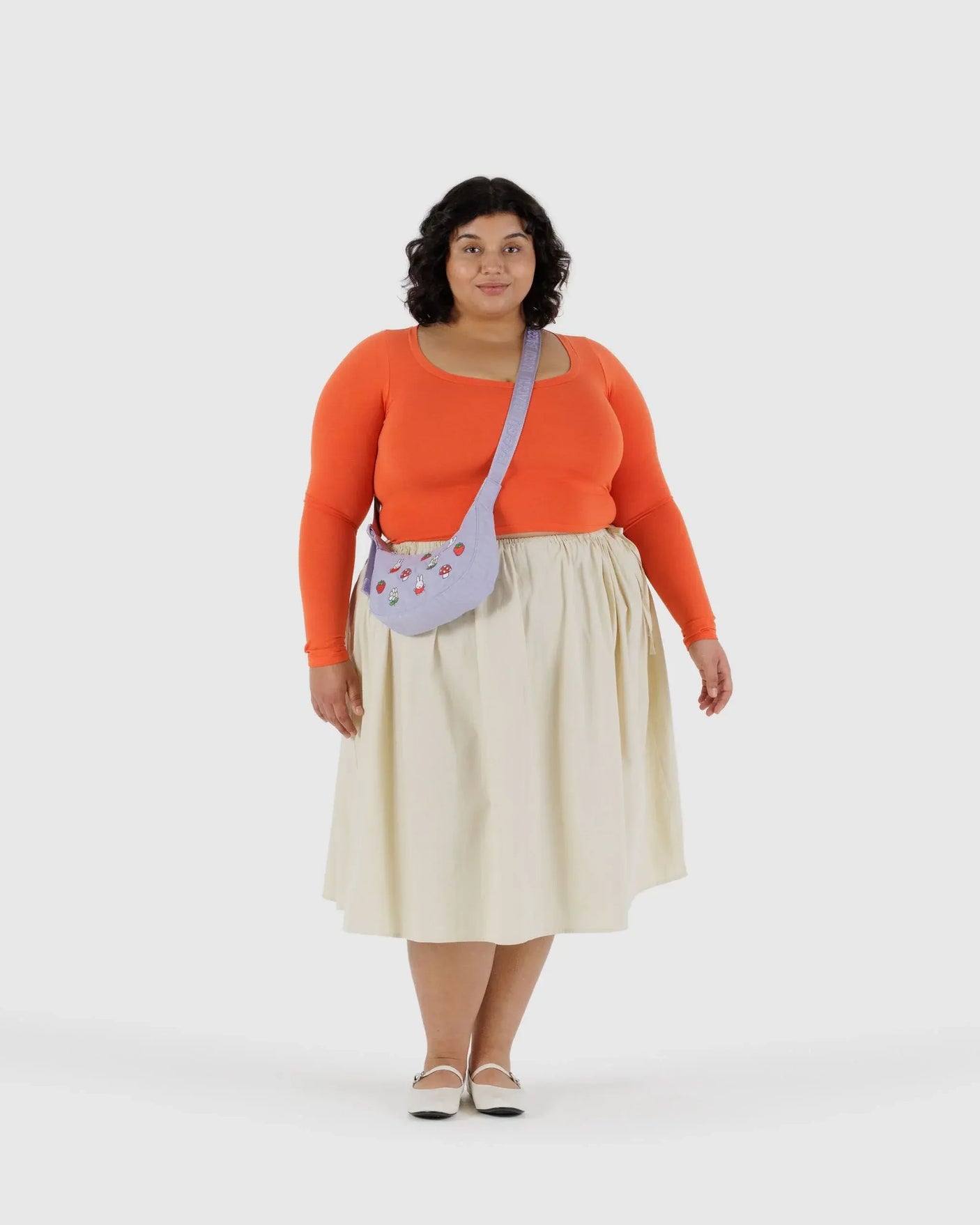 A woman faces forward, wearing an orange long-sleeve top, cream skirt, white shoes, and carries the Baggu Miffy x Baggu Small Nylon Crescent bag in light purple, featuring charming Miffy embroidery.