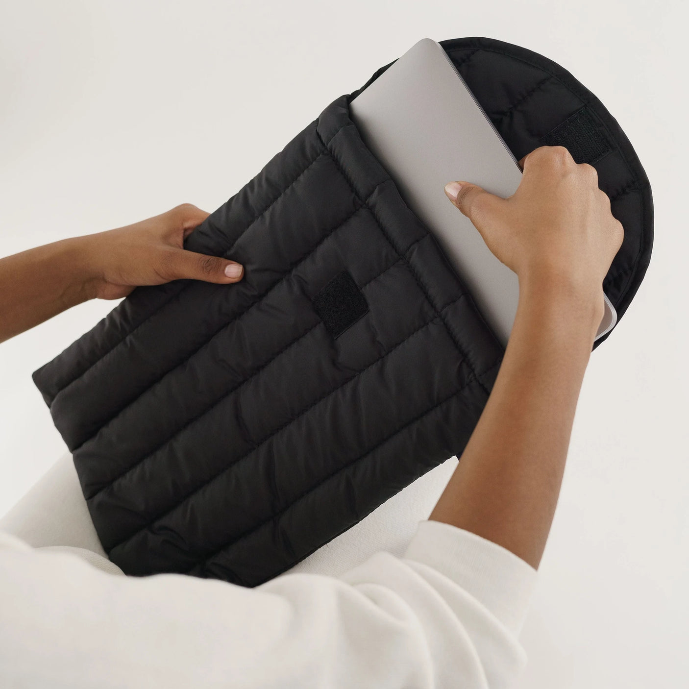 A person gently places a laptop into the Baggu Puffy Laptop Sleeve 16" in Black, made from recycled nylon and providing padded protection against the plain background.