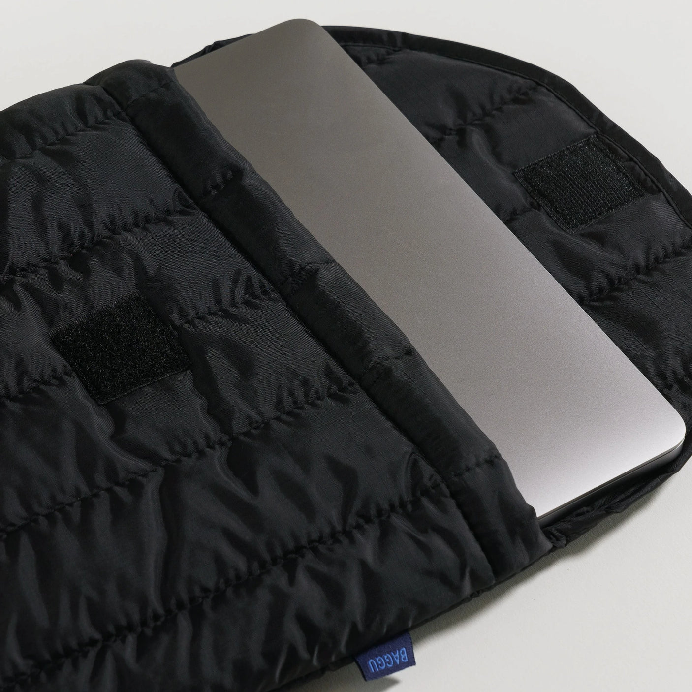 A Baggu Puffy Laptop Sleeve 16" in black, made from recycled nylon, partially slides out to reveal a laptop resting elegantly on a white surface.