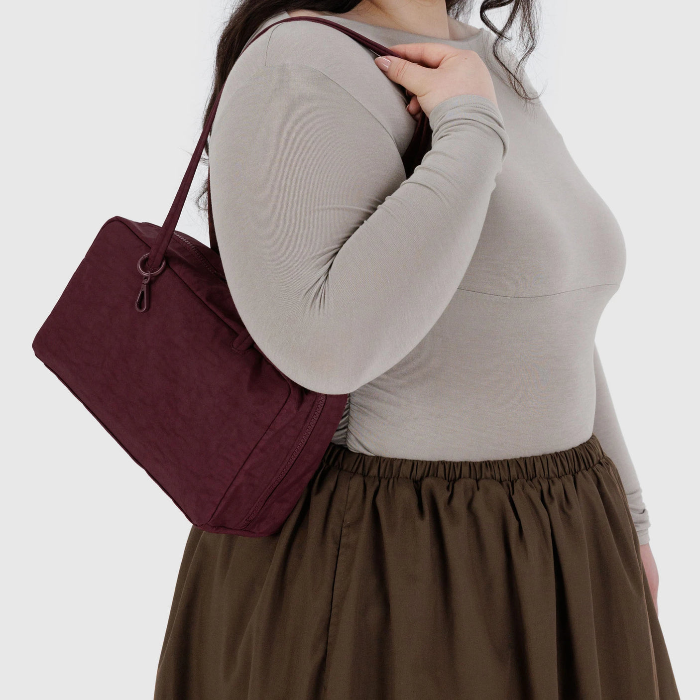 A person in a beige long-sleeve top and brown skirt holds a Baggu Nylon Bowler Bag - Mahogany by Baggu against a plain light background.