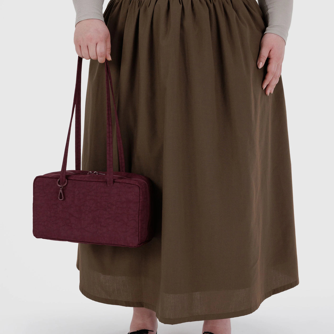 A person in a long brown skirt holds the Baggu Nylon Bowler Bag - Mahogany by Baggu, featuring two handles.