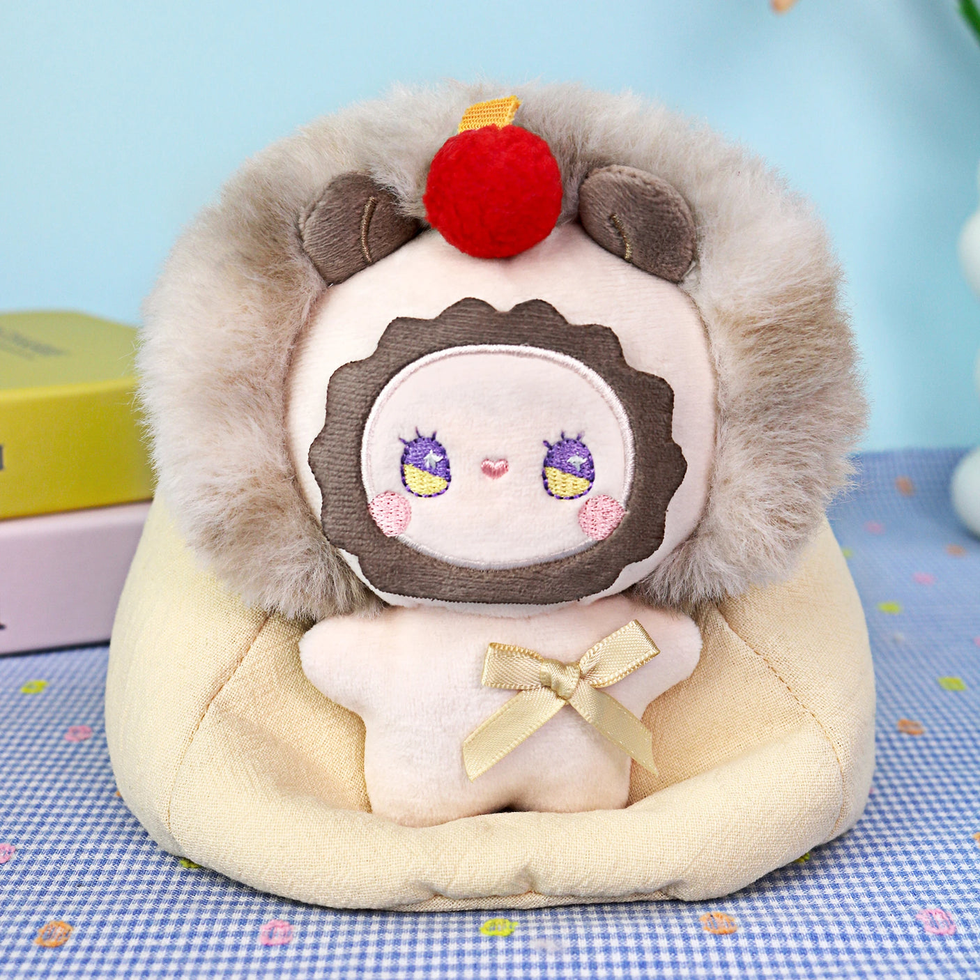 A plush toy with a round face, faux fur hood, and red pom-pom sits on a cream cushion with a yellow bow—the Emma Fuzzy Family Bag Charm - Blind Box by Lucky Emma, an adorable animal bag charm perfect for collectors.