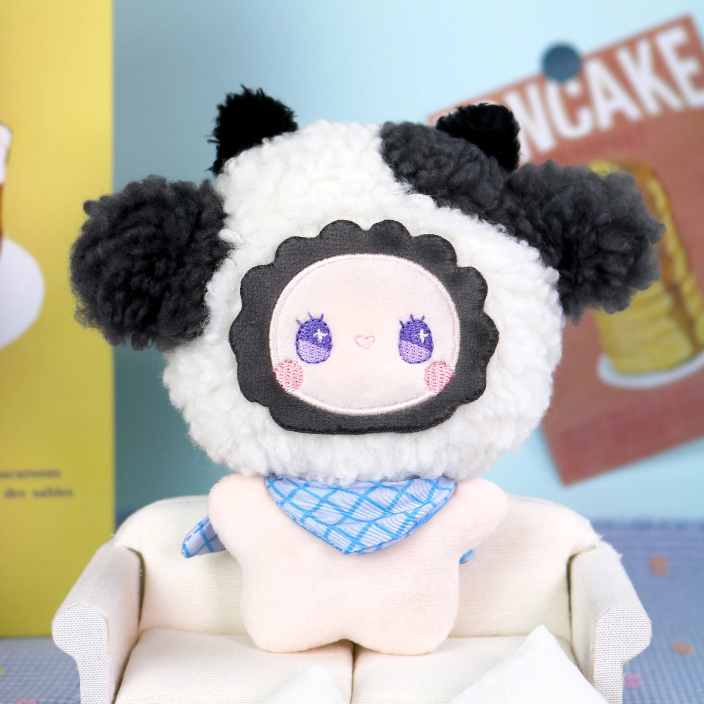 The Emma Fuzzy Family Keychain Blind Box by Lucky Emma features a plush doll in a sheep costume with black ears and a blue scarf, seated on a white sofa—ideal as a keychain or bag charm. Bright wall décor enhances the scene’s cute appeal.