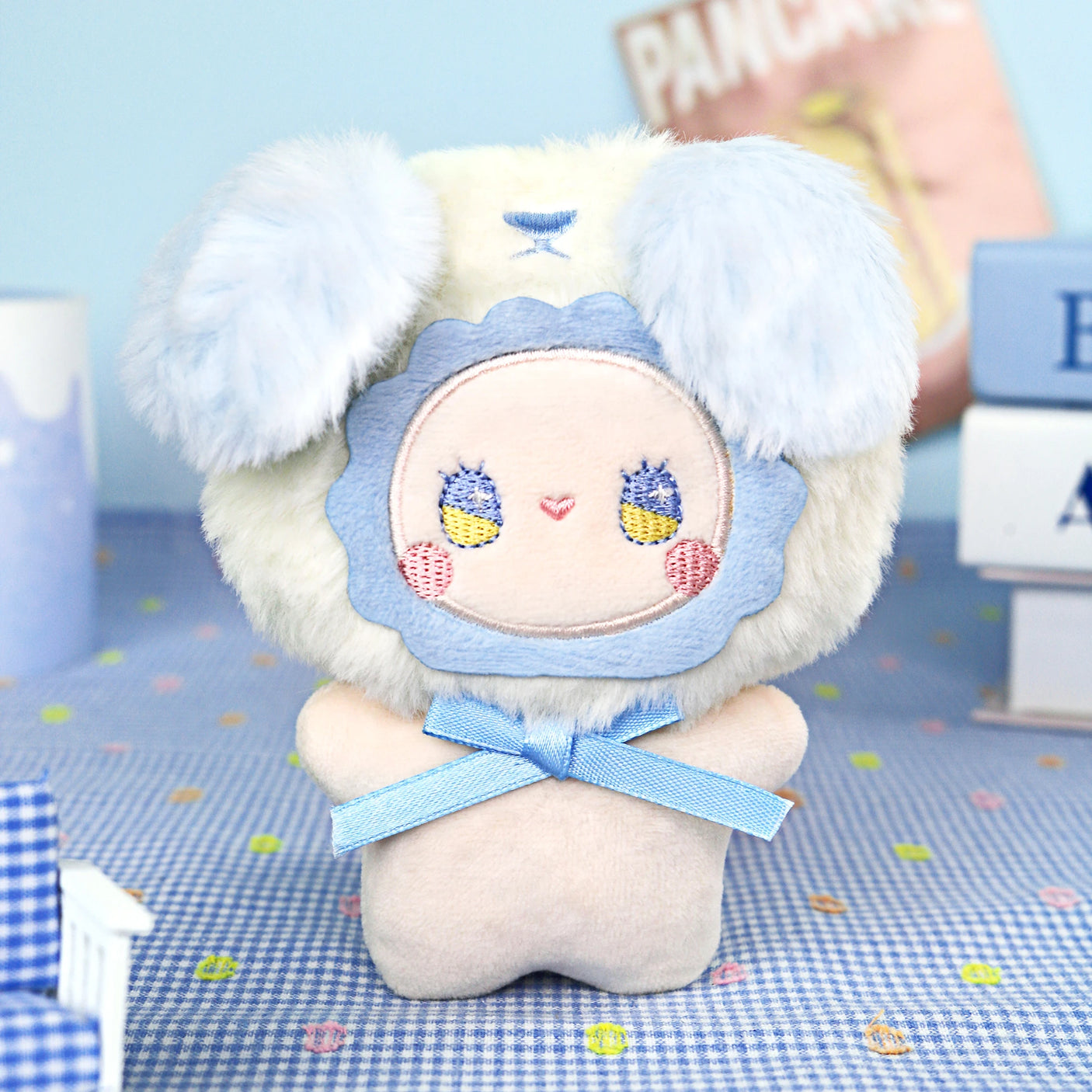 The Lucky Emma "Emma Fuzzy Family Keychain Blind Box" features a plush toy with a round embroidered face, blue ears, and a blue ribbon—perfect as a cute keychain or bag charm. Books and soft props set the scene in the background.