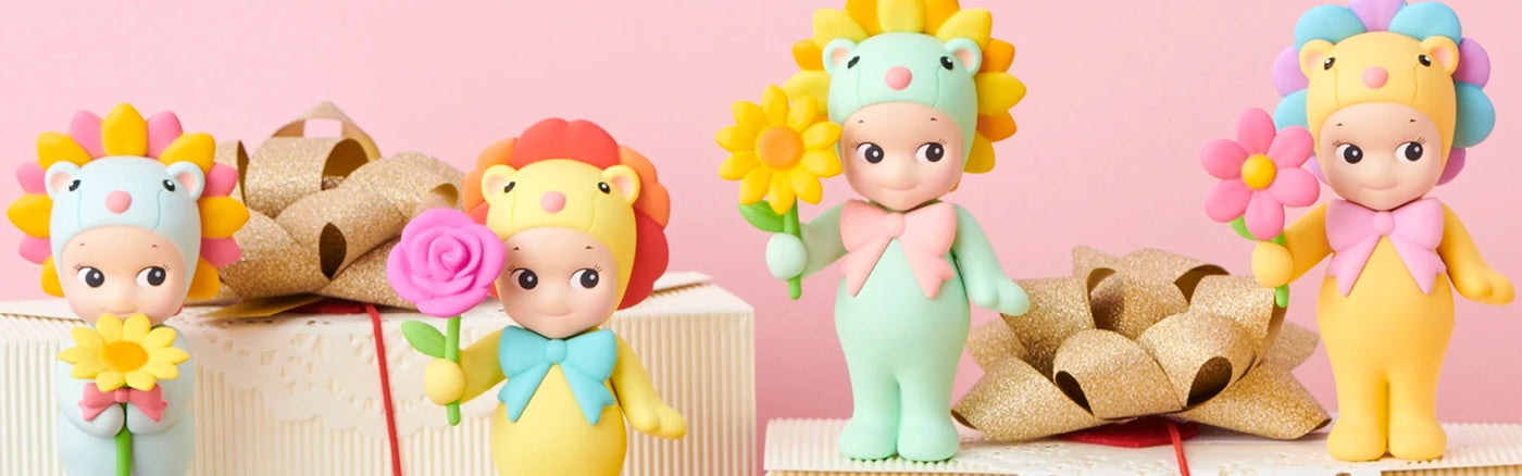 Four small figurines dressed in colorful animal-themed costumes holding flowers with gift boxes and gold bows on a pink background