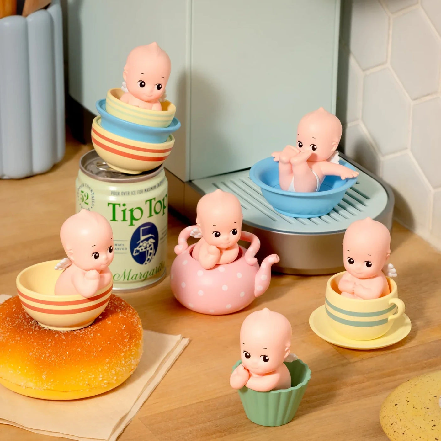 Small bald baby dolls sitting inside various colorful ceramic cups, bowls, and a pink polka-dot teapot on wooden countertop
