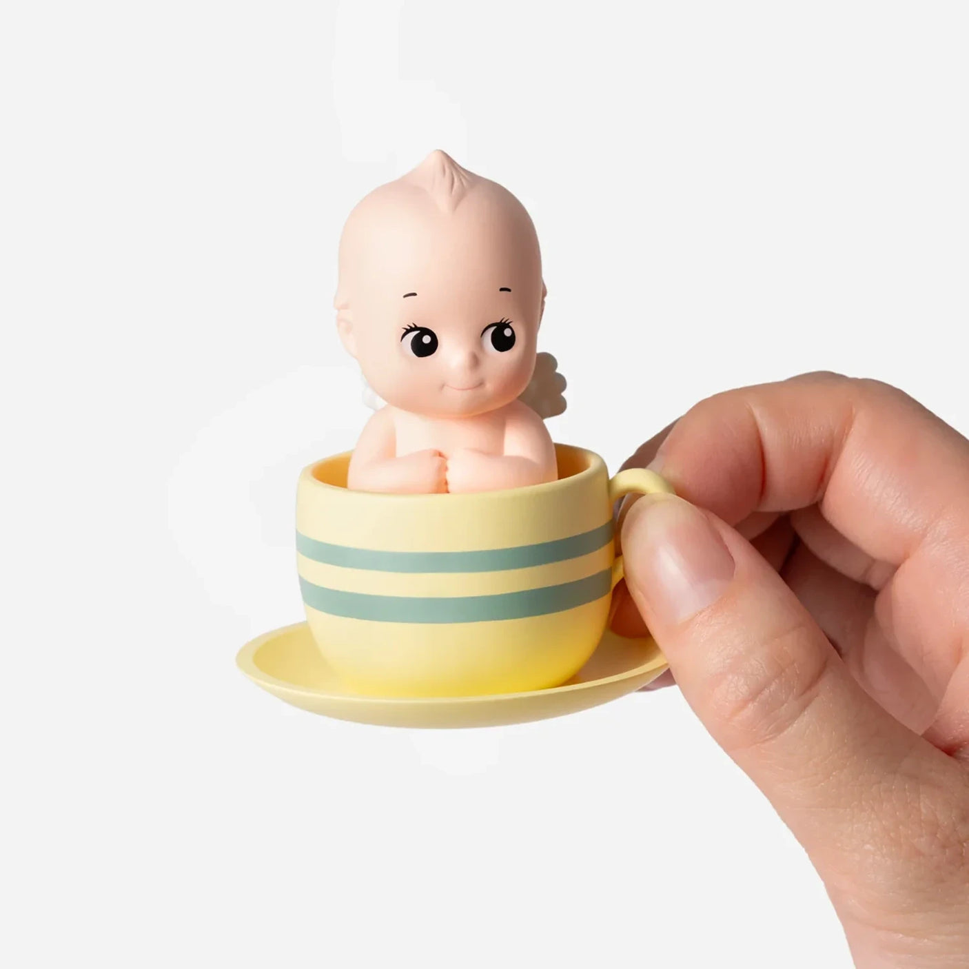 Hand holding small doll with angel wings sitting inside yellow and green striped teacup on matching saucer