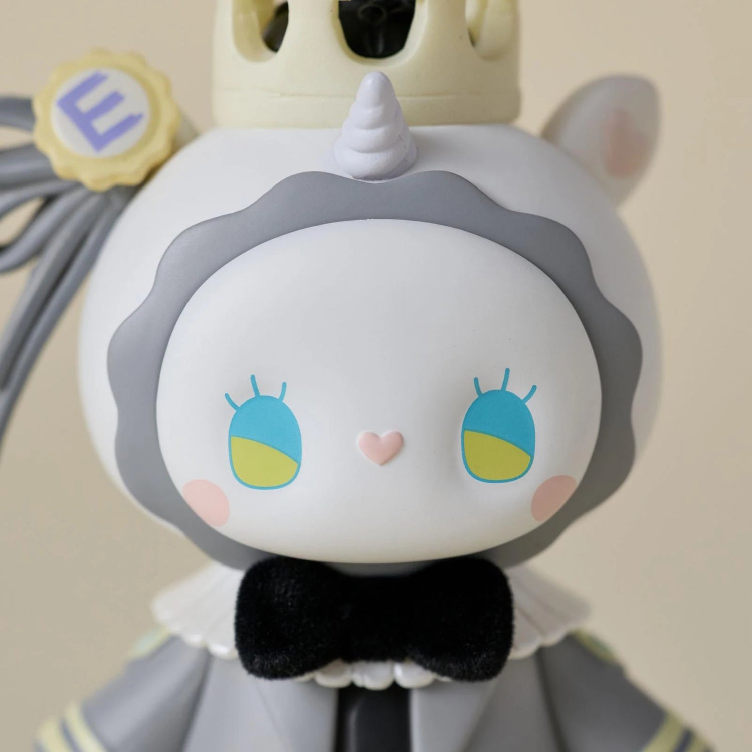 A close-up of a cute, cartoon-like figurine with a unicorn horn, crown, big blue-and-yellow eyes, and black bow tie in gray and white—this is a Secret Figure from the Lucky EMMA Secret Forest Wedding Blind Box by Lucky Emma.