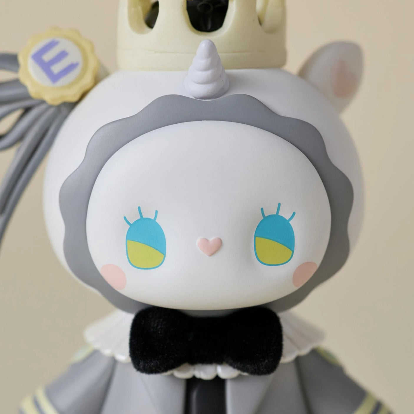 A close-up of a cute, cartoon-like figurine with a unicorn horn, crown, big blue-and-yellow eyes, and black bow tie in gray and white—this is a Secret Figure from the Lucky EMMA Secret Forest Wedding Blind Box by Lucky Emma.