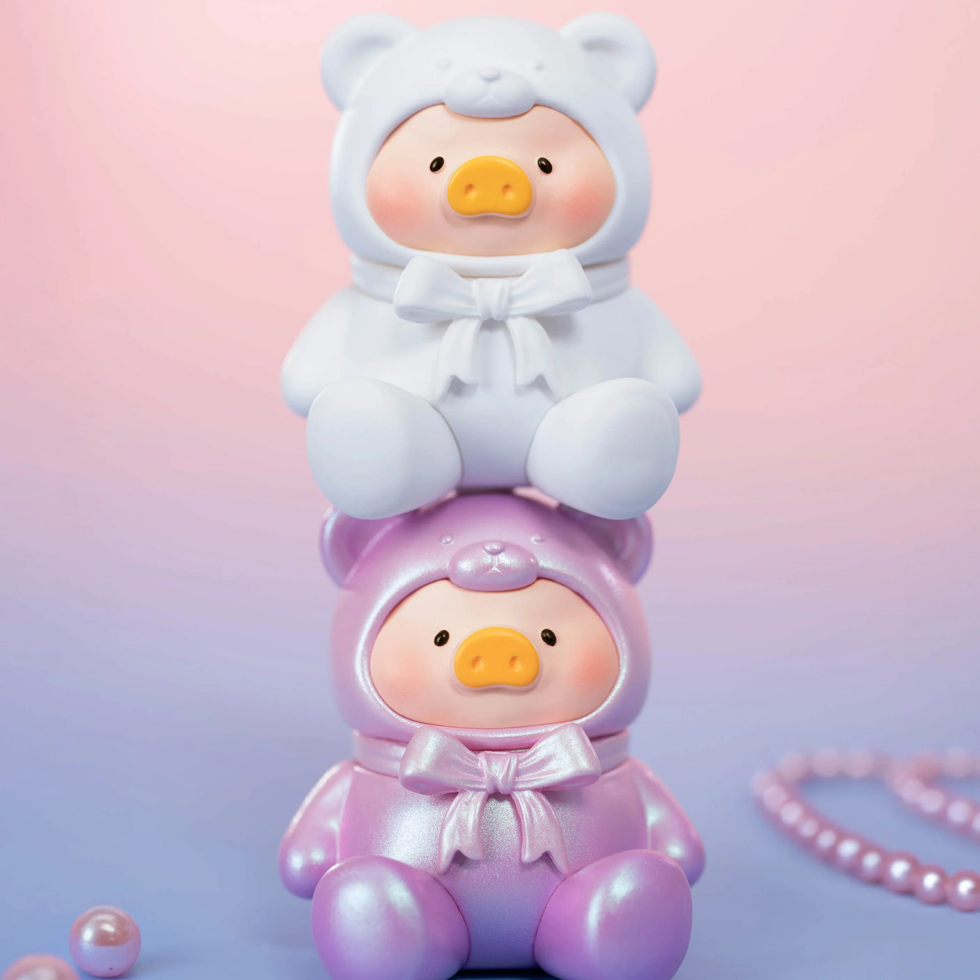 Two LuLu The Piggy - Mini Teddy Blind Bag figures, by LuLu The Piggy, in white and pink bear costumes are stacked together on a pink-purple gradient background, with shimmering pearls adding extra charm to the scene.