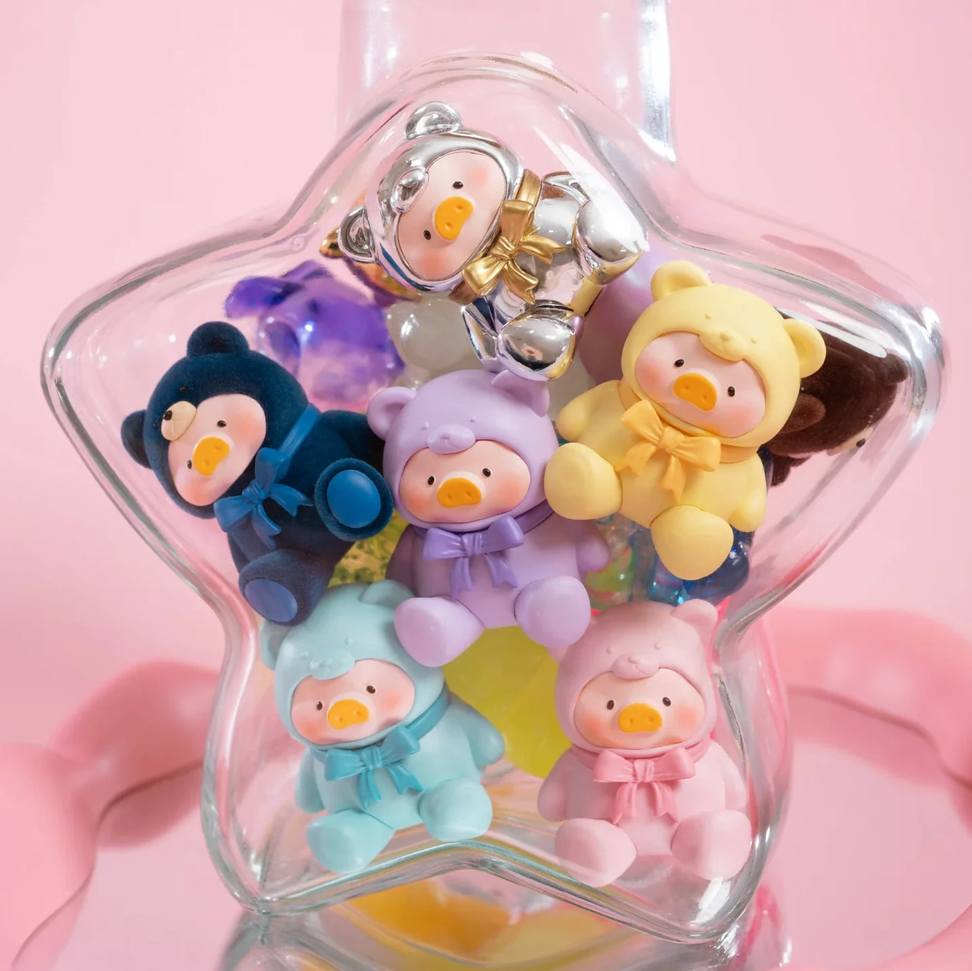 Six small, pastel-colored LuLu The Piggy figurines in bear onesies are displayed in a star-shaped jar, resembling the LuLu The Piggy - Mini Teddy Blind Bag collectible, with a pink background.