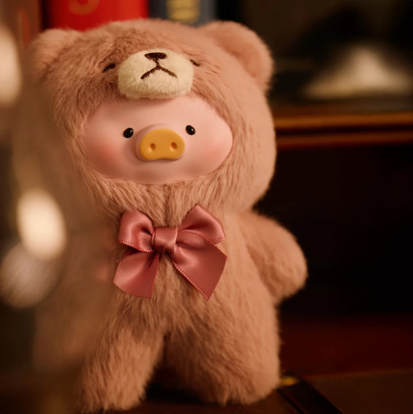 Teddy LuLu the Piggy Plush Pal - Blind Box by LuLu The Piggy stands adorably in a teddy bear onesie with a bow, posed in front of a blurred bookshelf.