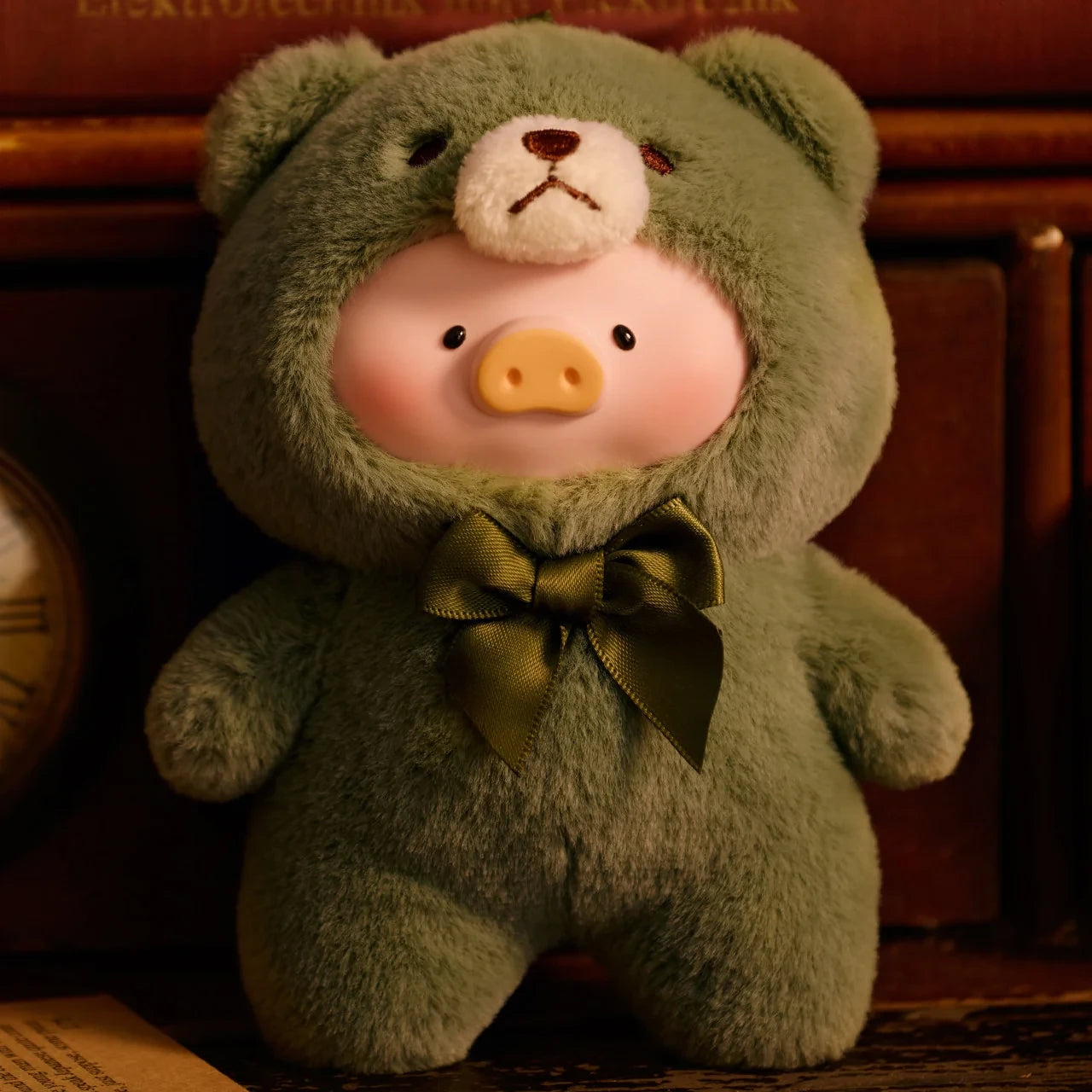A LuLu The Piggy Teddy LuLu the Piggy Plush Pal - Blind Box wears a green bear onesie with a bow tie, posed before wooden shelves and books.