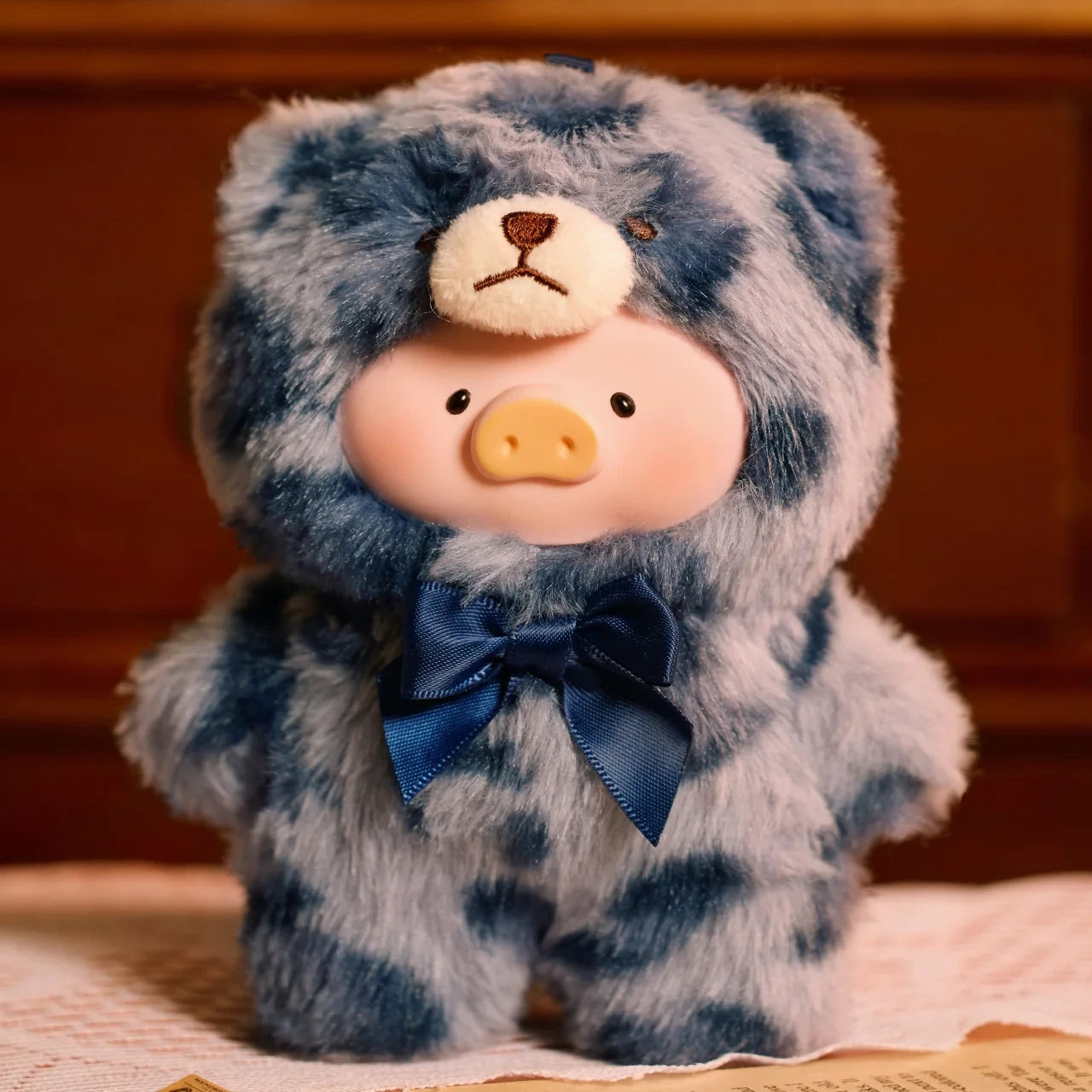 The LuLu The Piggy "Teddy LuLu the Piggy Plush Pal - Blind Box" features a piggy plush in a blue, bear-patterned teddy bear onesie with a matching bow tie, displayed on a patterned indoor surface.