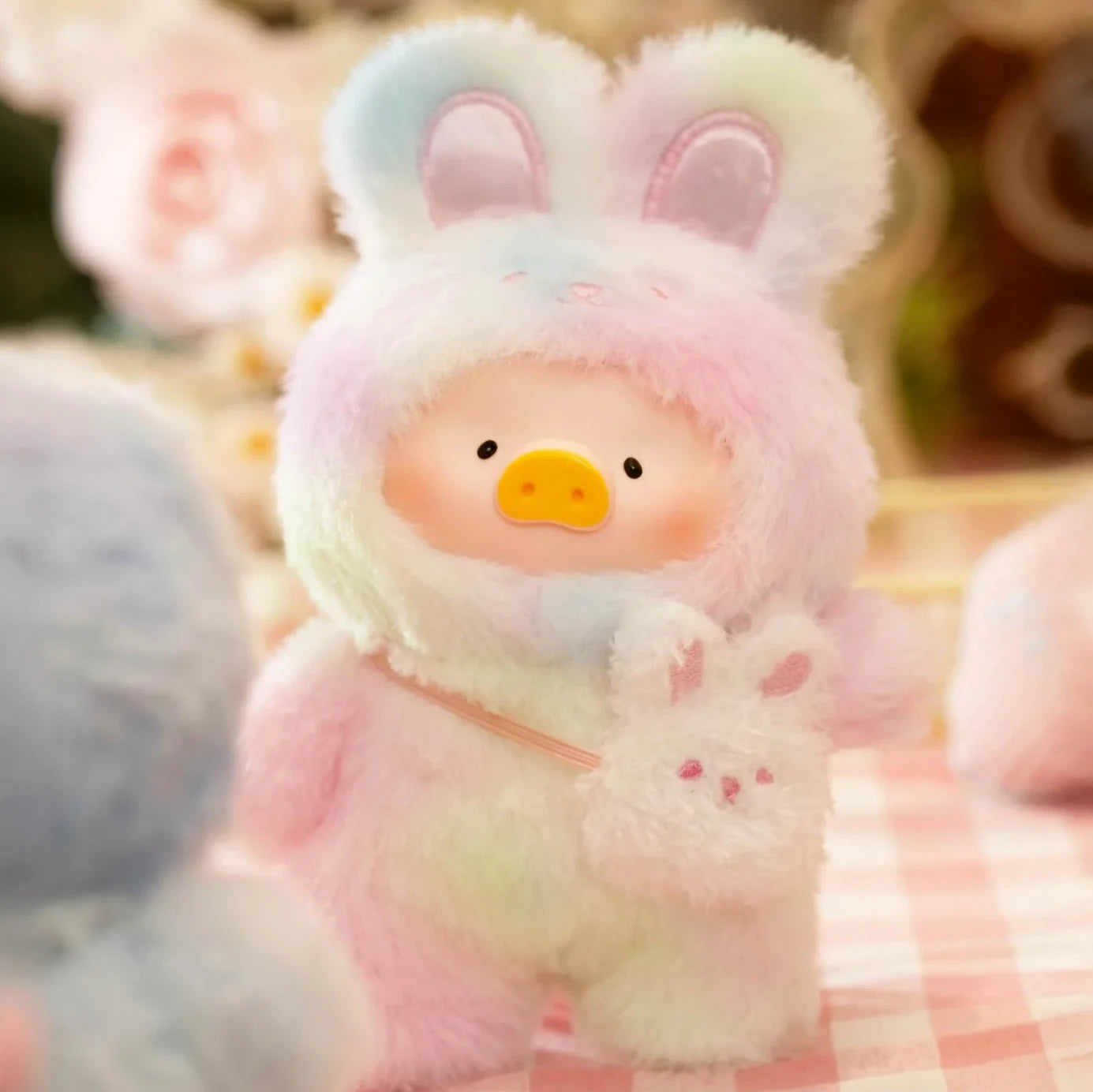 Joyful Time Lulu The Piggy Plush Pal - Blind Box by LuLu The Piggy features a charming plush in a pastel rainbow bunny costume with a duck face, perfect as a collectible keychain or gift. Displayed on a pink checkered surface with blurred floral decor.