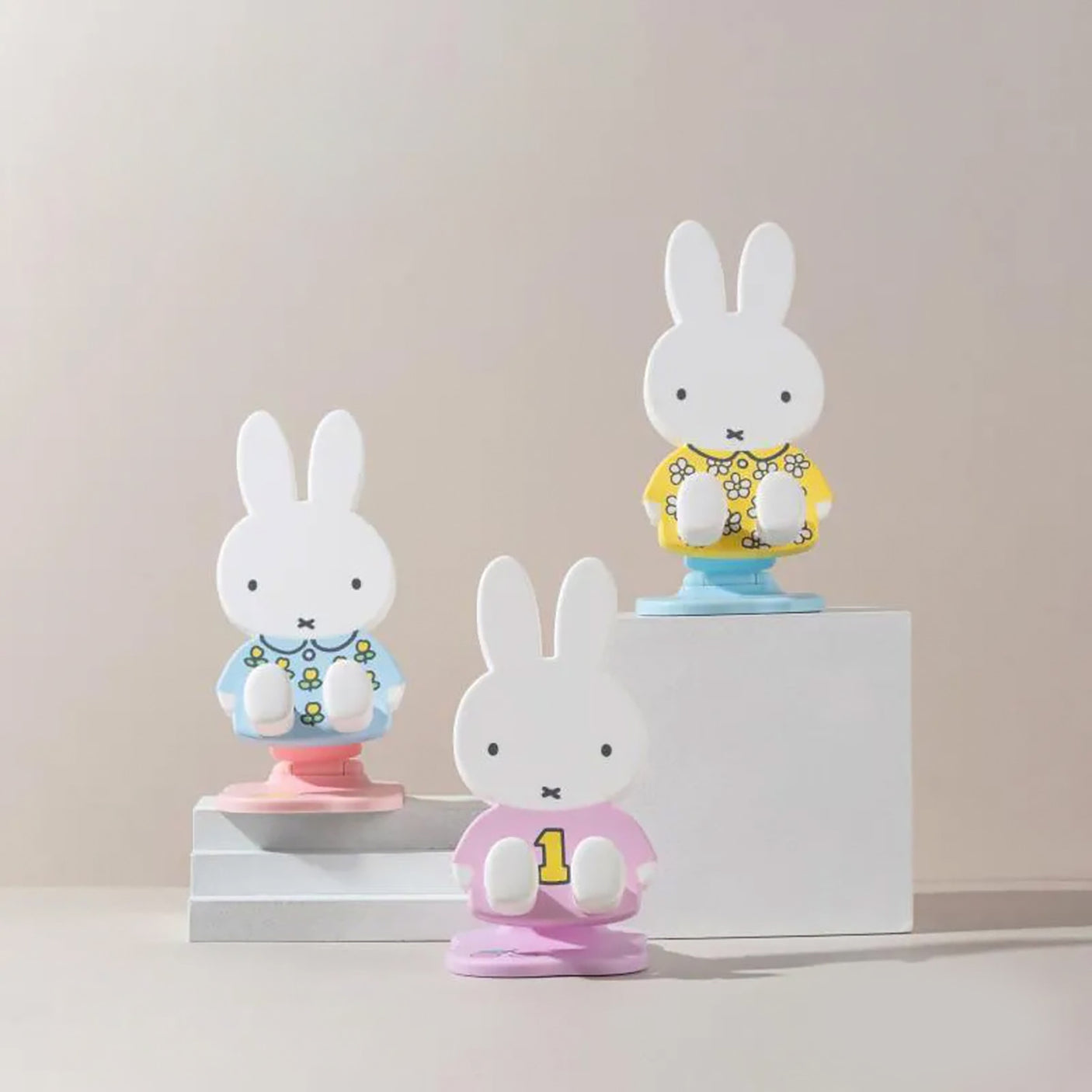 Three Miffy phone and tablet stands in pastel pink, blue, and yellow outfits displayed on white blocks against neutral background