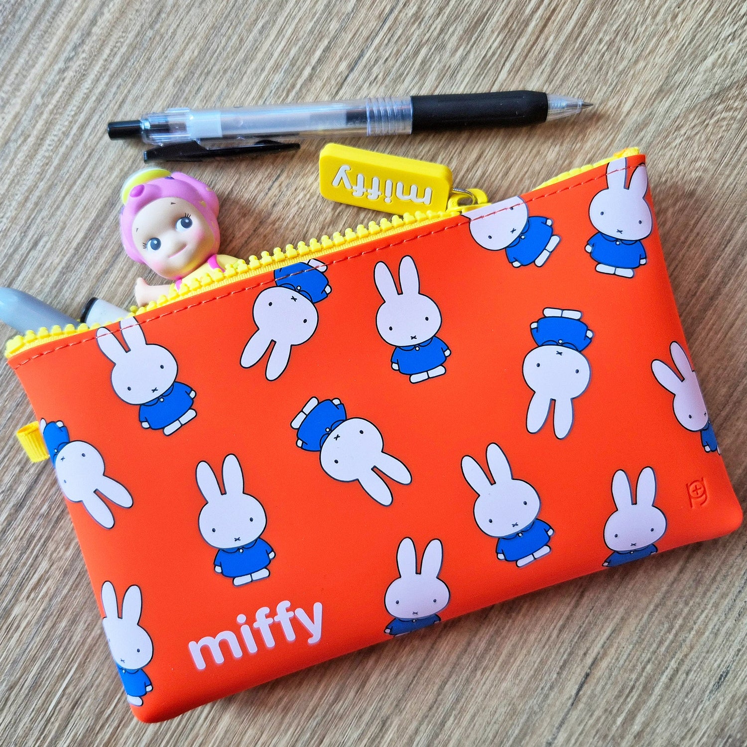 A Miffy Bunny Pattern Case 19cm, along with a pen and a small doll, rests on the wooden surface.