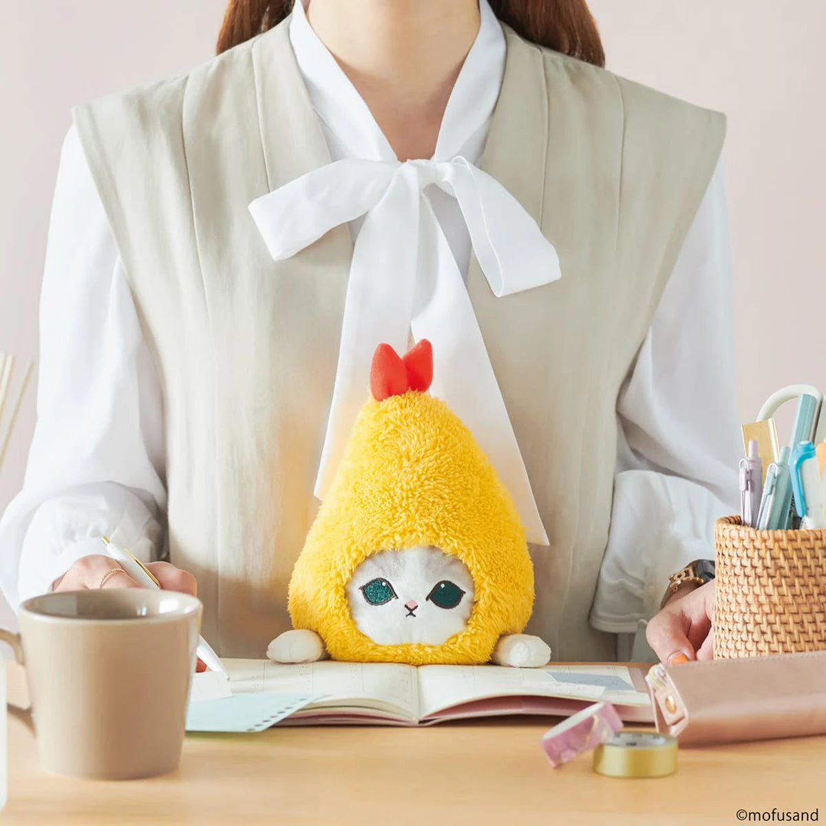 A person sits at a desk with a Mofusand Posture Plush Buddy - Fried Shrimp pen holder, an open notebook, cup, and assorted stationery items.