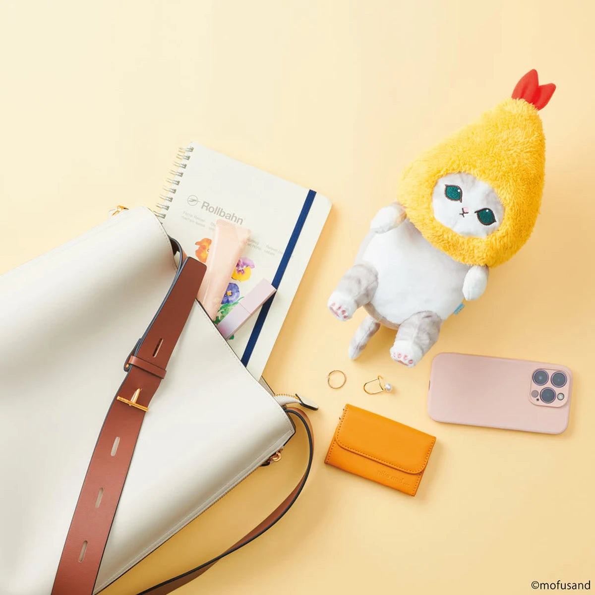 A handbag, notebook, smartphone, ring, earrings, small orange wallet, and a cute Mofusand Posture Plush Buddy - Fried Shrimp by Mofusand are arranged on a light yellow surface.