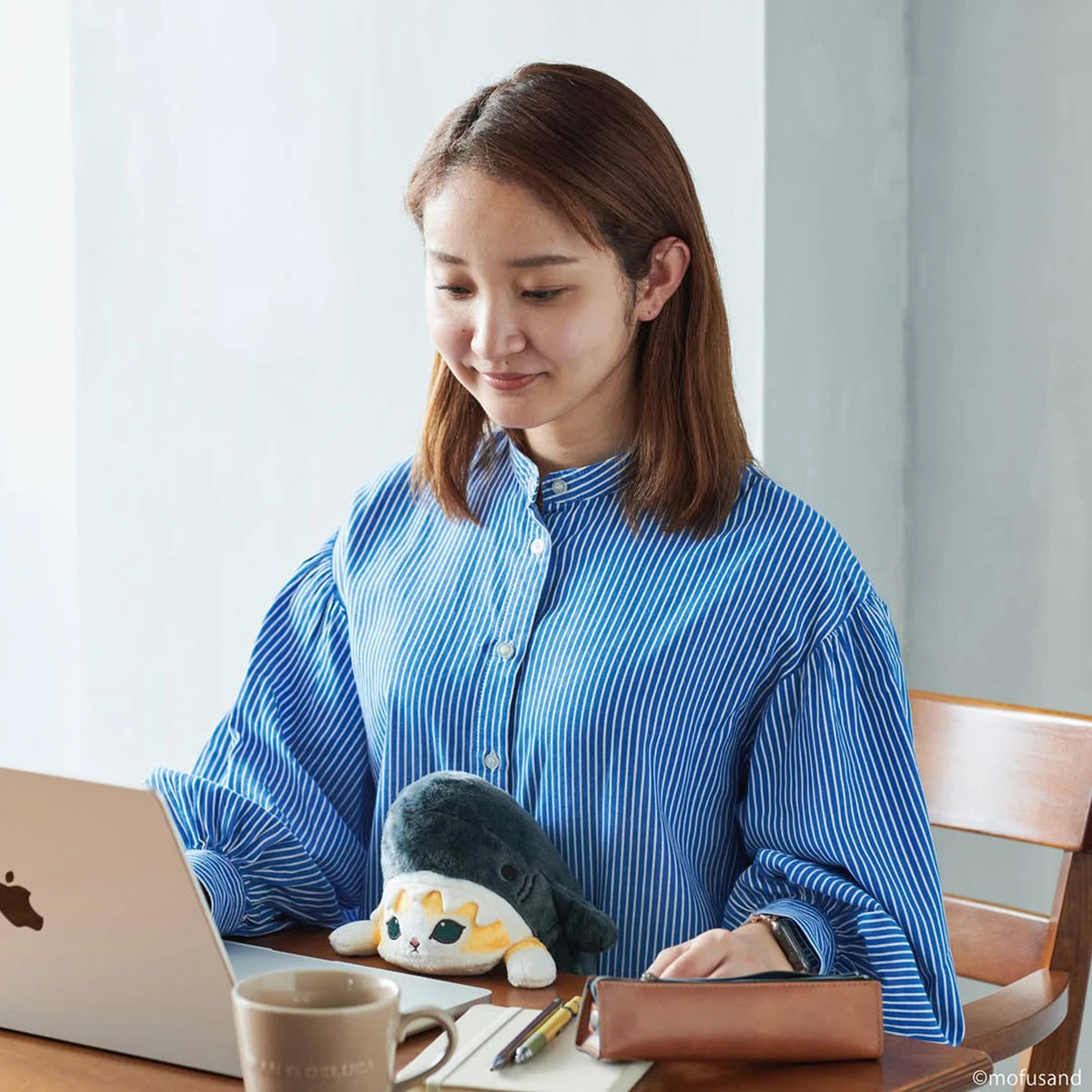 A woman in a blue striped shirt works on a laptop at a table with her Mofusand Posture Plush Buddy - Shark, along with a mug, wallet, and phone.