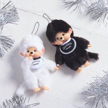 Two Monchhichi keychain dolls—a Monchhichi Colours Bag Charm in white by Monchhichi and a brown one—lie on a white surface with silver tinsel, making adorable gifts or cute bag charms.