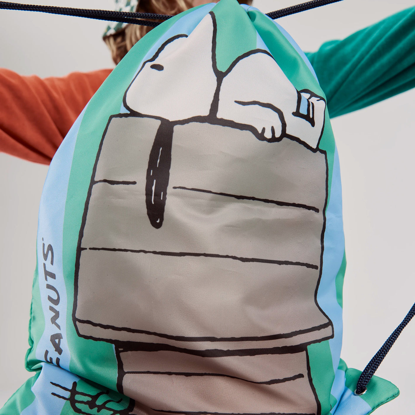 Light blue and green Peanuts drawstring bag showcasing Snoopy relaxing on his doghouse with black cords and Peanuts text on side
