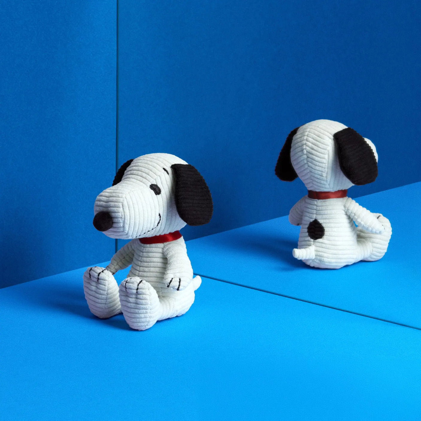 Two cream corduroy Snoopy plush toys sitting, black ears and red collar, side view