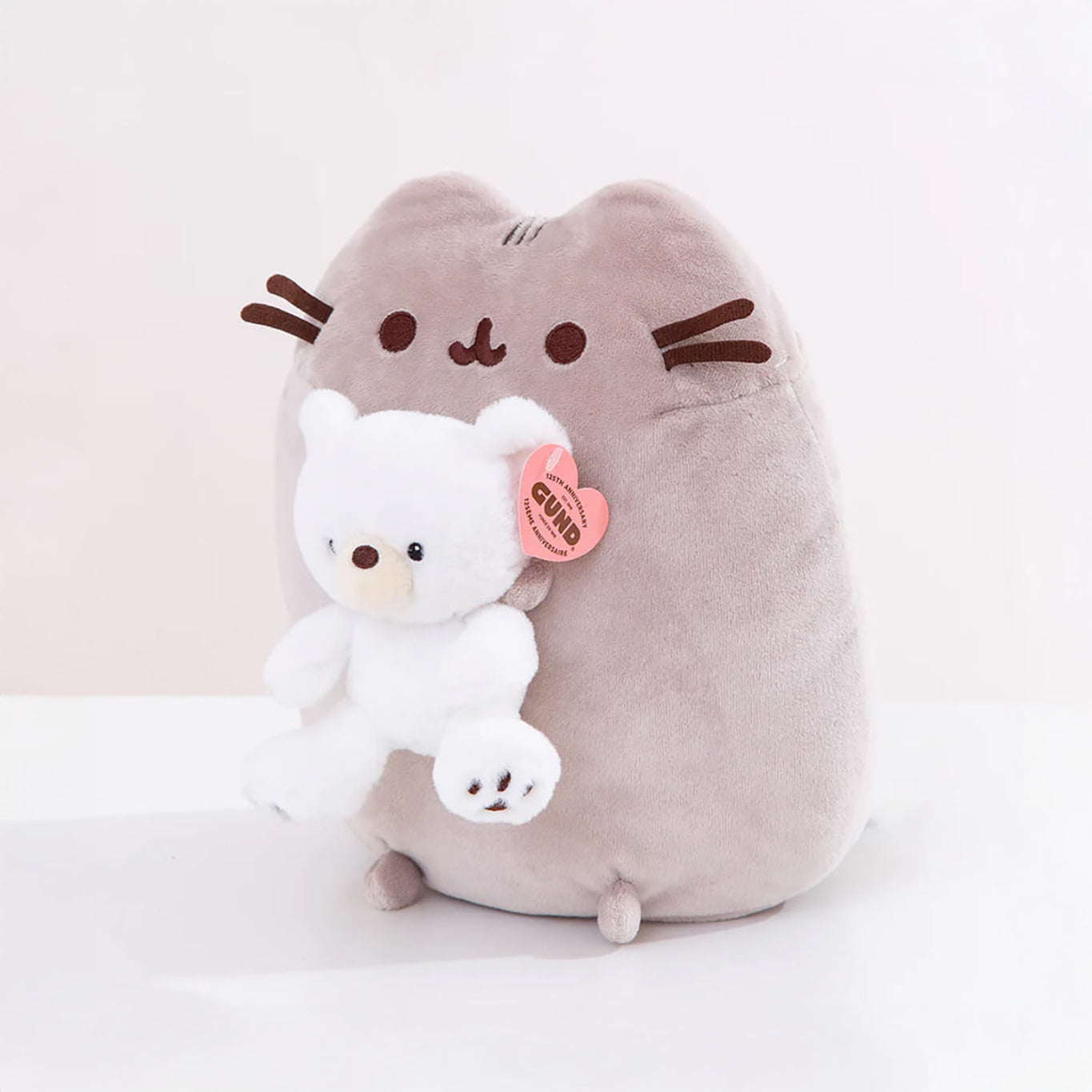 A plush gray Pusheen toy lovingly holds a small companion—an adorable white teddy bear, part of the limited edition Pusheen x GUND Kai Bear Plush collection.