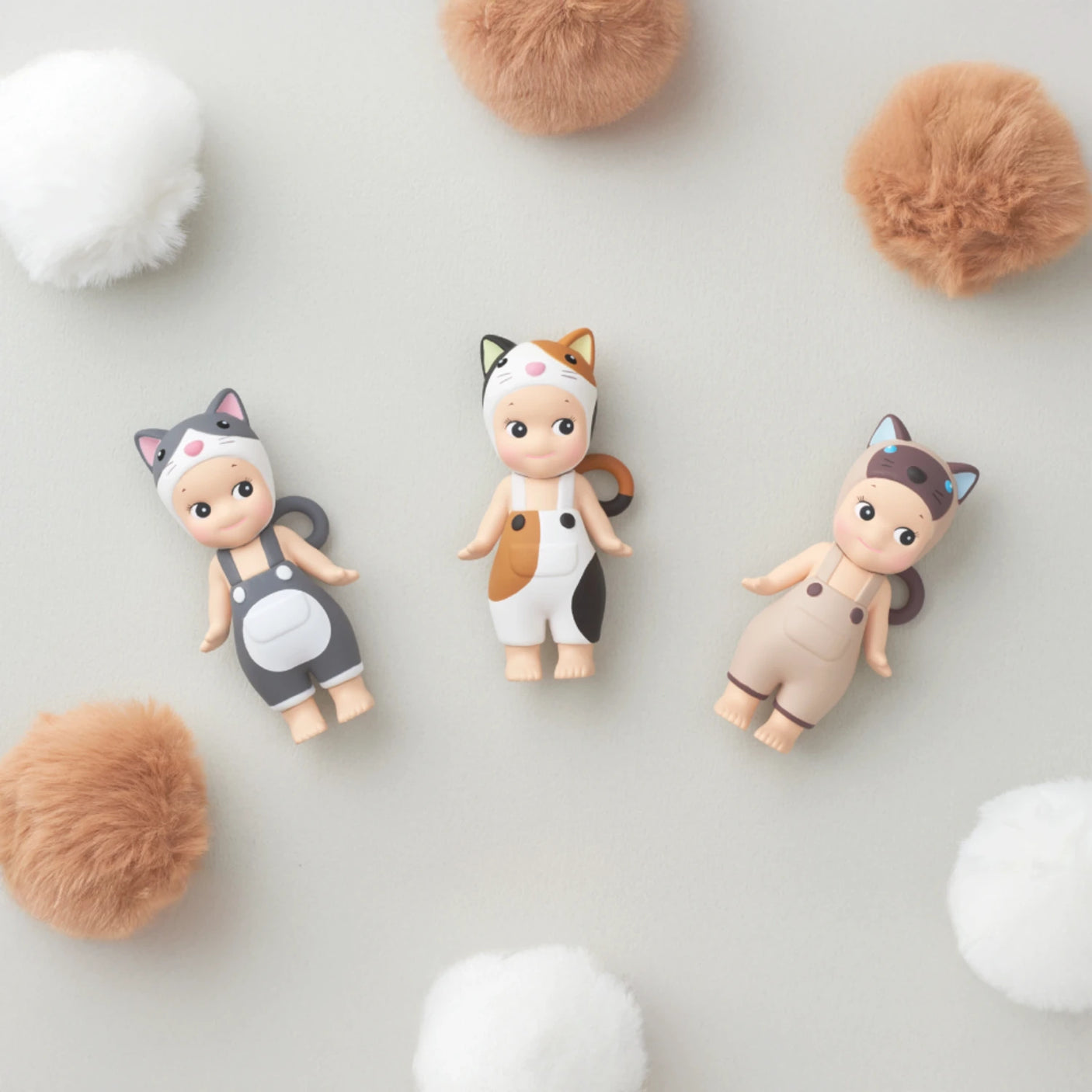 Three figures from the Sonny Angel Cat Life Series - Blind Box, under the Sonny Angel brand, are placed on a gray surface surrounded by fluffy white and brown pom-poms.