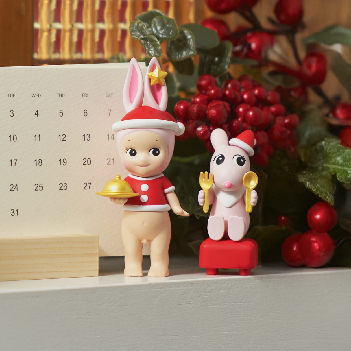 The Sonny Angel Christmas Dinner Blind Box, featuring a bunny-eared figure in a Santa outfit with a dish and a rabbit holding utensils, sits on a shelf with a calendar and red berry decor, creating the perfect scene for an enchanting Christmas dinner.