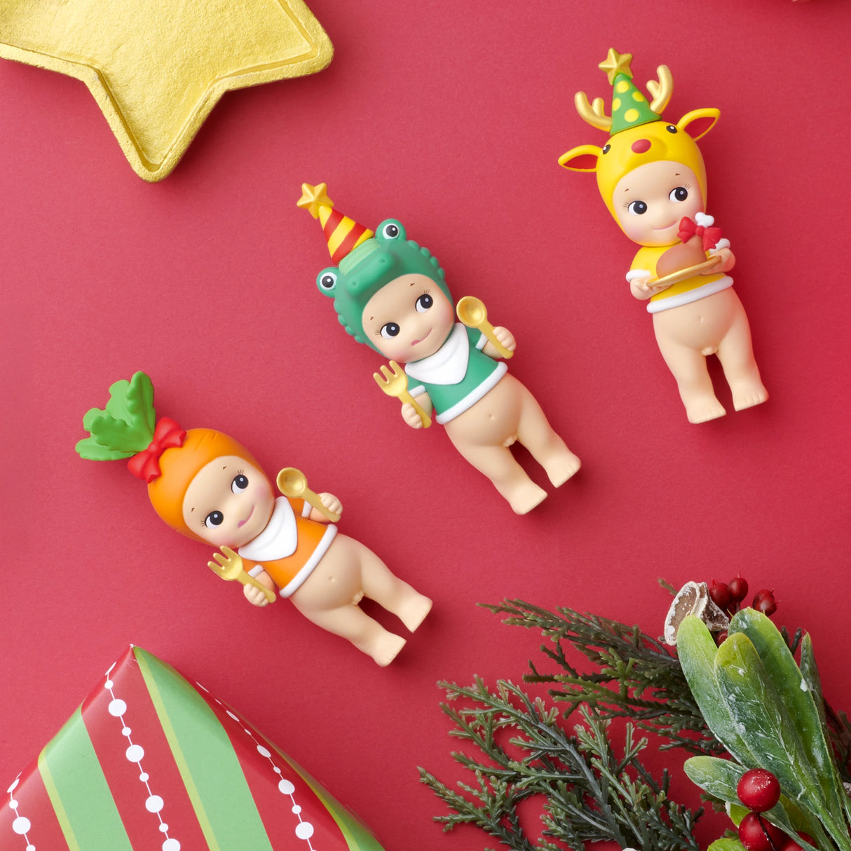 Three Sonny Angel figures from the Christmas Dinner - Blind Box collection are dressed in festive costumes and hats, each holding a spoon. They are set against a red backdrop decorated with a gold star and Christmas greenery, capturing the whimsical charm of a holiday dinner.
