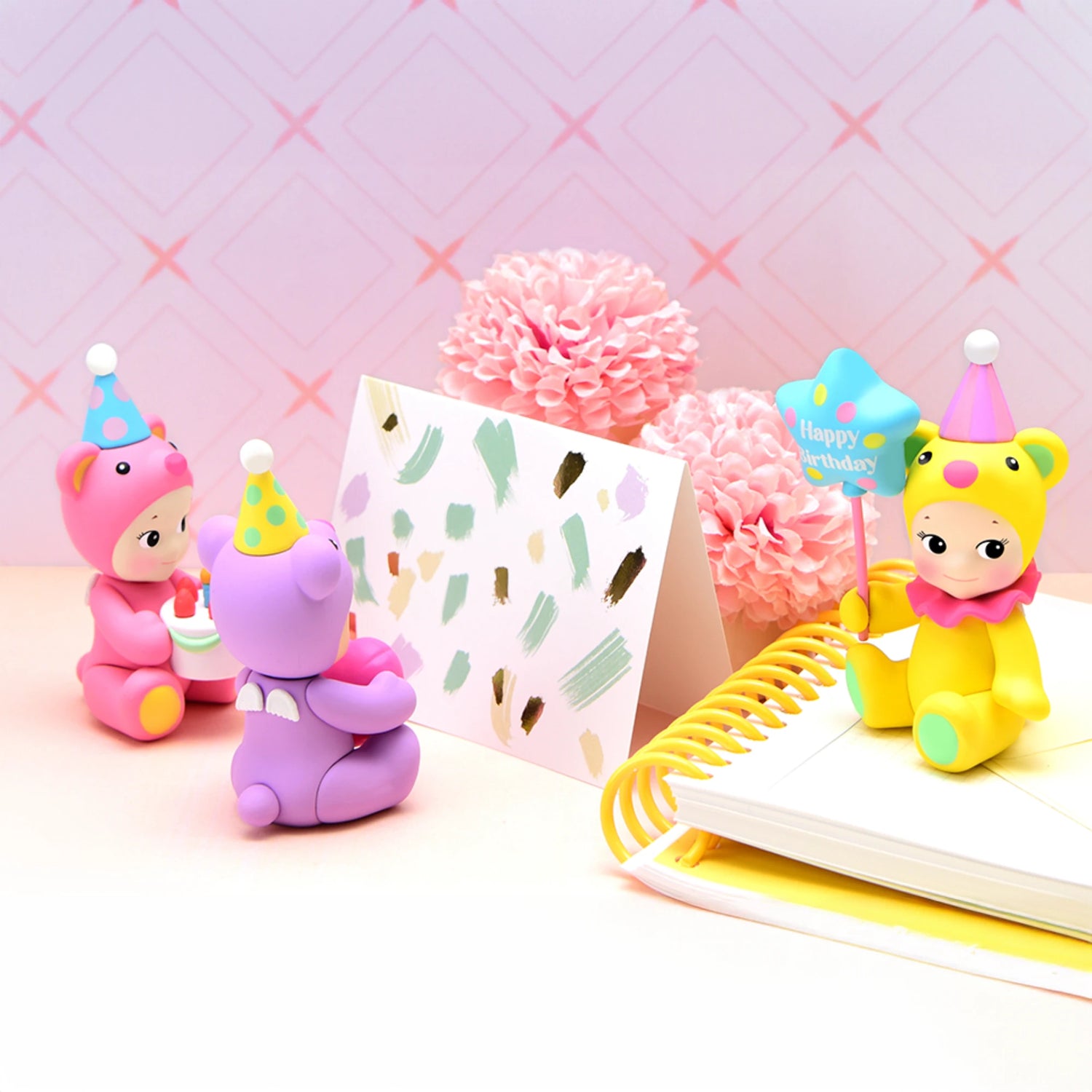 Three Sonny Angel Birthday Bear Gift figurines, adorned with party hats, are arranged near a card, pink flowers, and a notebook on a pastel background.