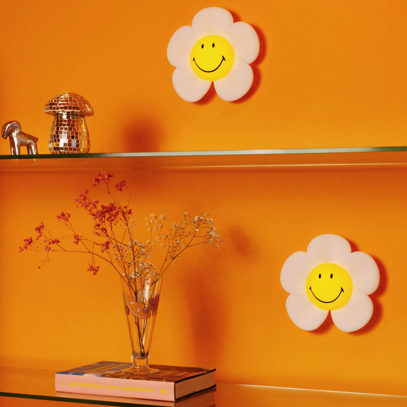 Two Mr Maria Smiley Daisy - Day & Night Lights are mounted on an orange wall above glass shelves with a vase of dried flowers, disco ball mushroom, small figurine, and a book, adding playful retro flower power charm.