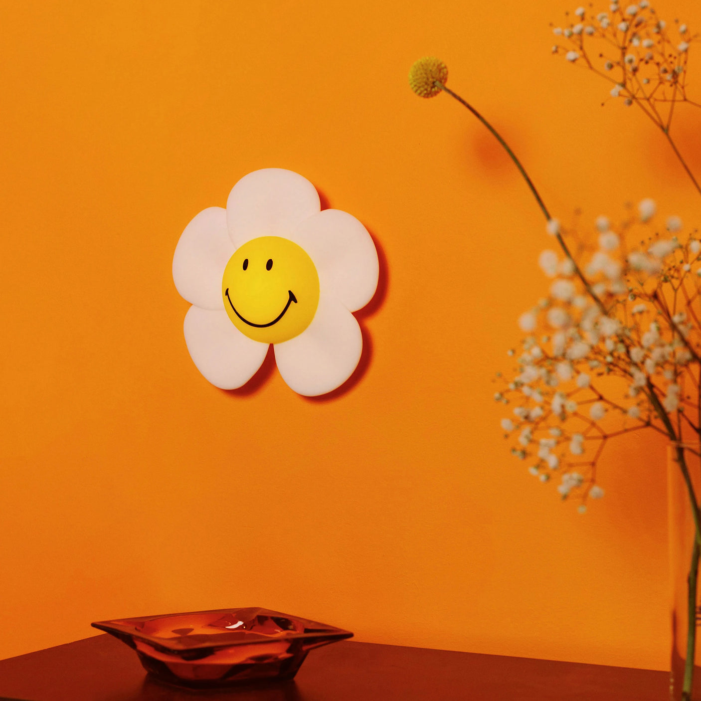 The Smiley Daisy - Day & Night Light by Mr Maria, featuring a cheerful face, decorates an orange wall above a dark surface with a metallic dish; dried flowers sit to the right.