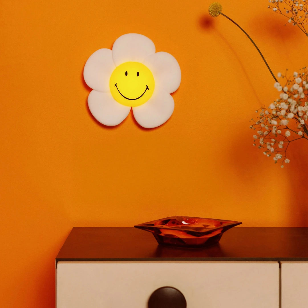 The Smiley Daisy - Day & Night Light by Mr Maria, featuring a cheerful daisy face, hangs above a cabinet with an orange glass dish, set against an orange wall decorated with colorful flowers.
