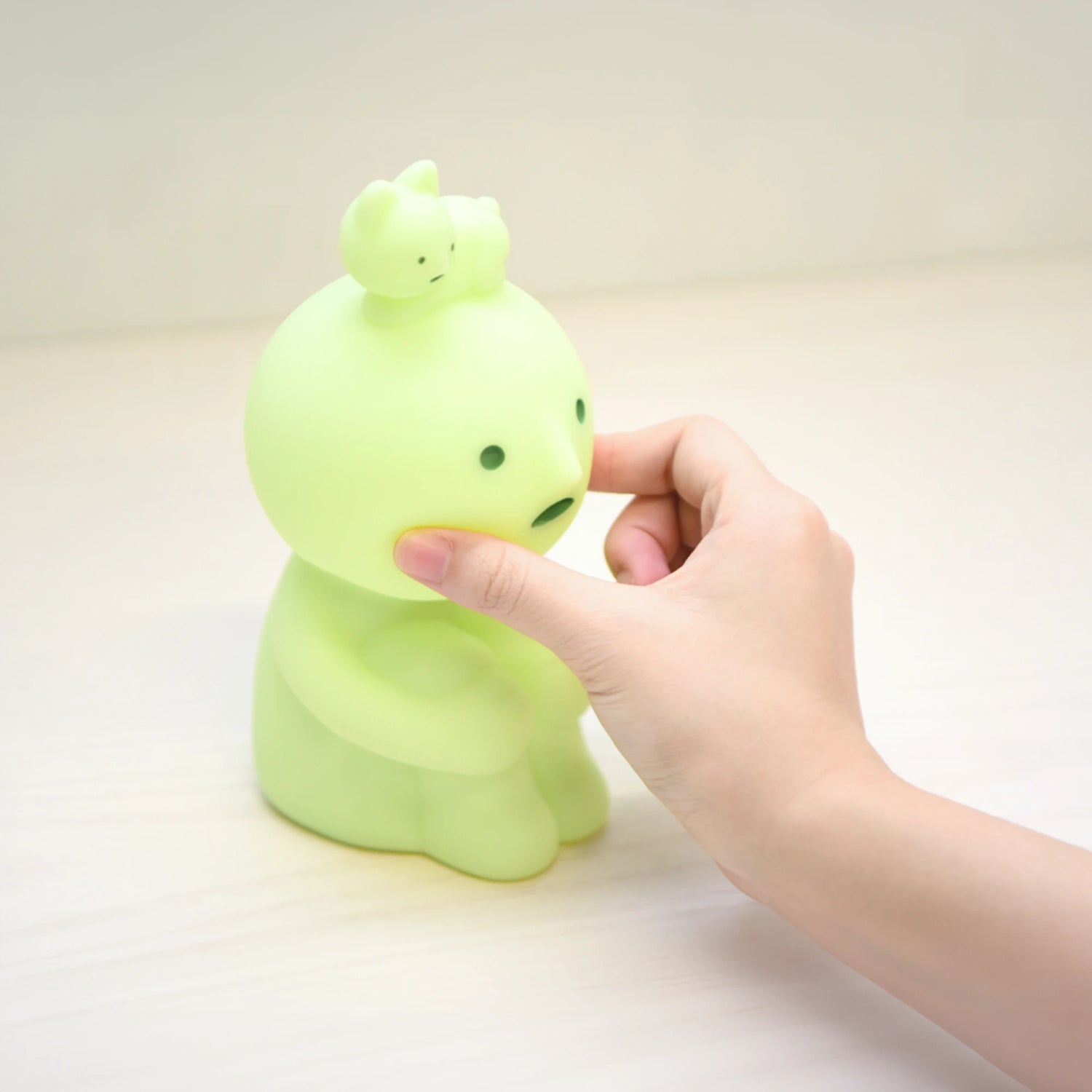 A hand squishes the cheek of a small, green, round-headed figurine that resembles a Smiski Touch Light nightlight by Smiski, with a smaller figure perched on its head, placed on a light surface.