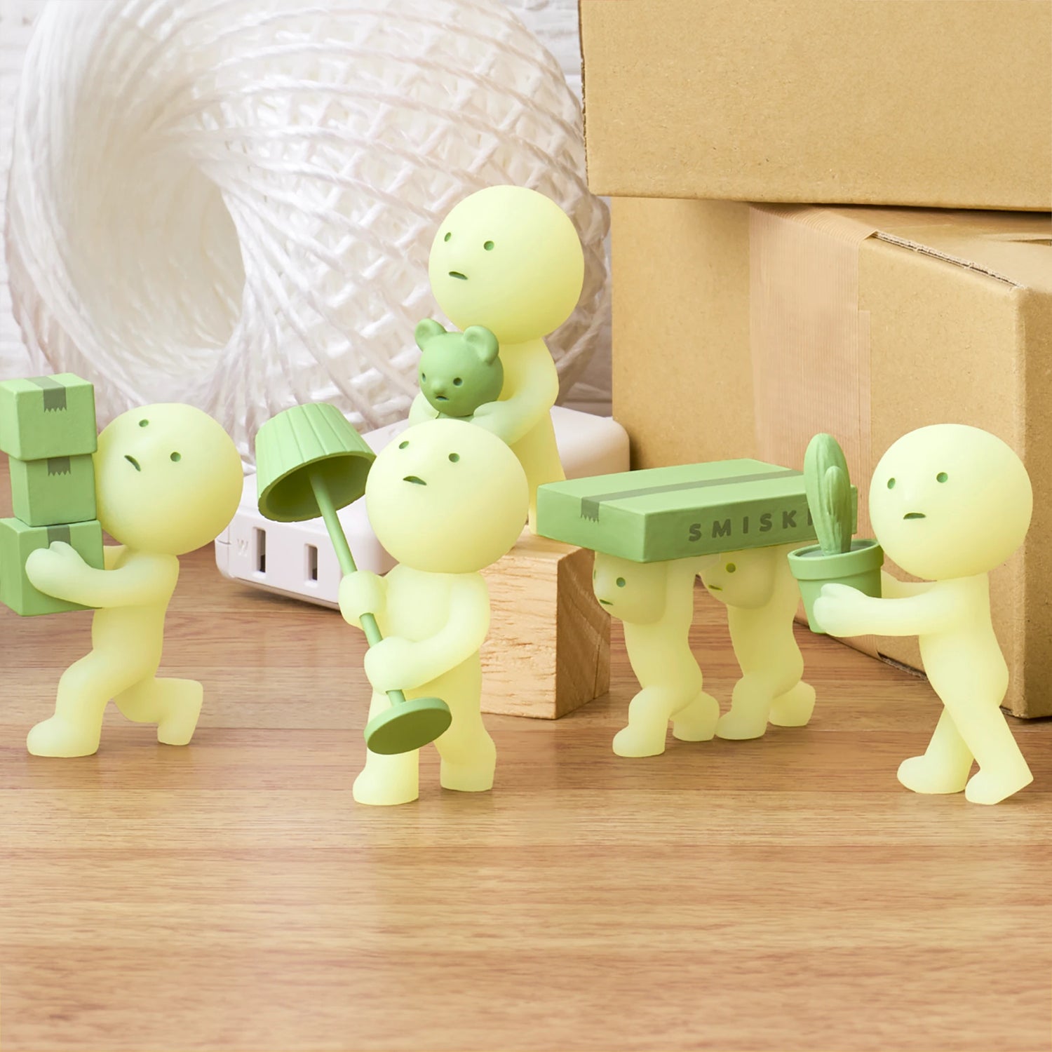 The Smiski Moving Series - Blind Box by Smiski features five small, cartoonish figures in pale green engaging in various activities such as carrying boxes and holding a plant, with one sporting a lampshade. These glow-in-the-dark collectibles are displayed against a wooden surface and cardboard boxes, making them the perfect addition to any toy collection.