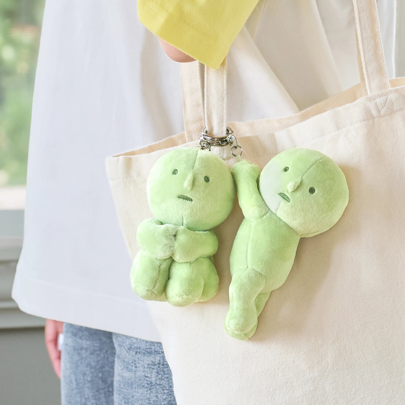 Two light green Smiski plush keychains attached to beige tote bag handles with silver keyrings