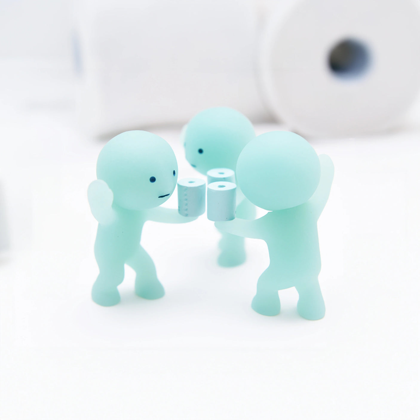 A trio of diminutive, light green glow-in-the-dark figures from the Smiski Toilet Series - Blind Box by Smiski hold tiny toilet paper rolls, positioned on a white surface with larger rolls in the background. Ideal for fans of whimsical mini collectible figures.
