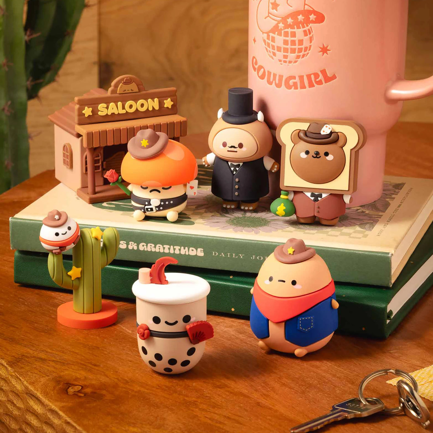 Collection of small vinyl figures including cowboy characters, a cactus, a bubble tea cup, and a miniature saloon building on books and wood surface