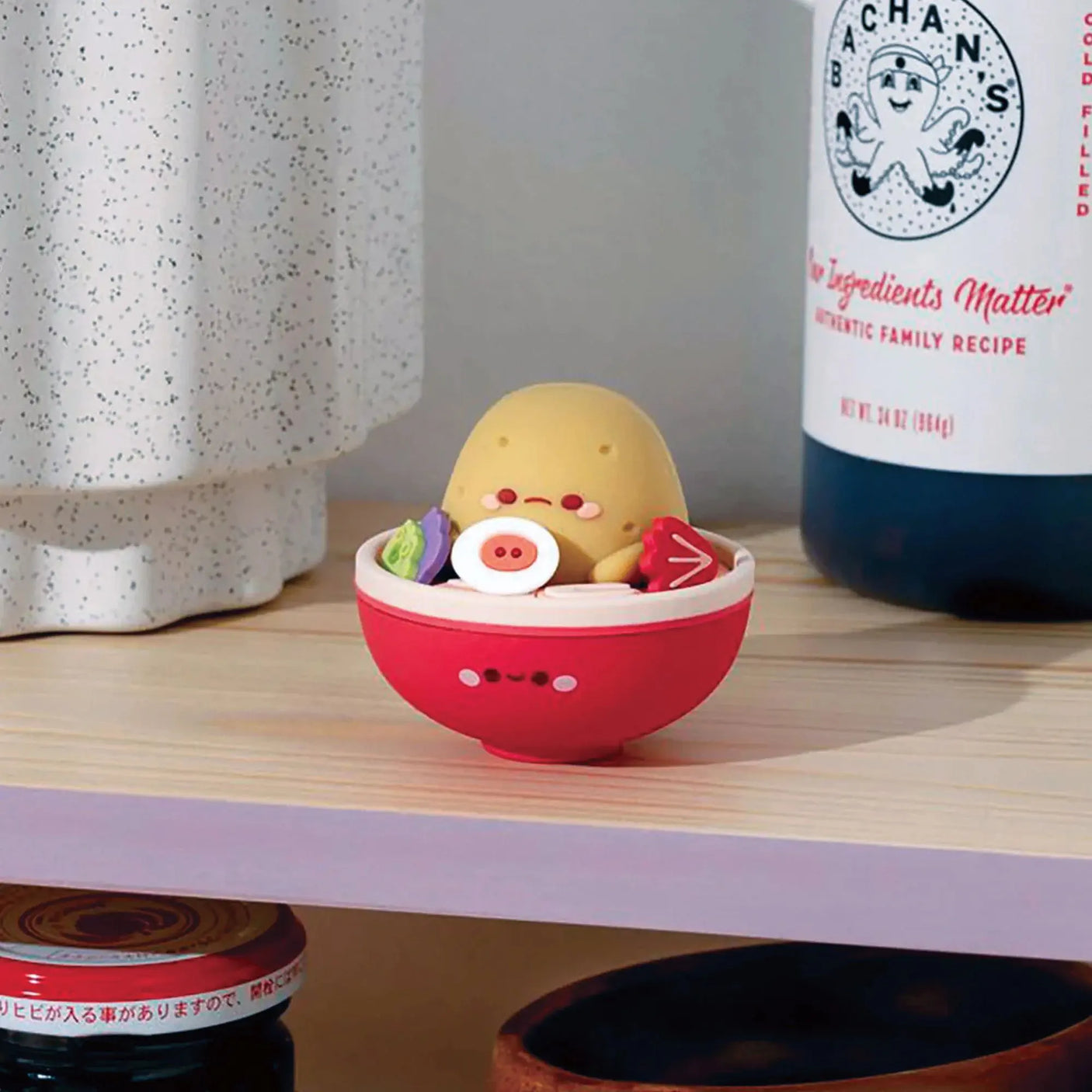 A vinyl figurine resembling a potato sits in a red bowl with tiny vegetables, placed on a shelf next to a speckled jar and a
