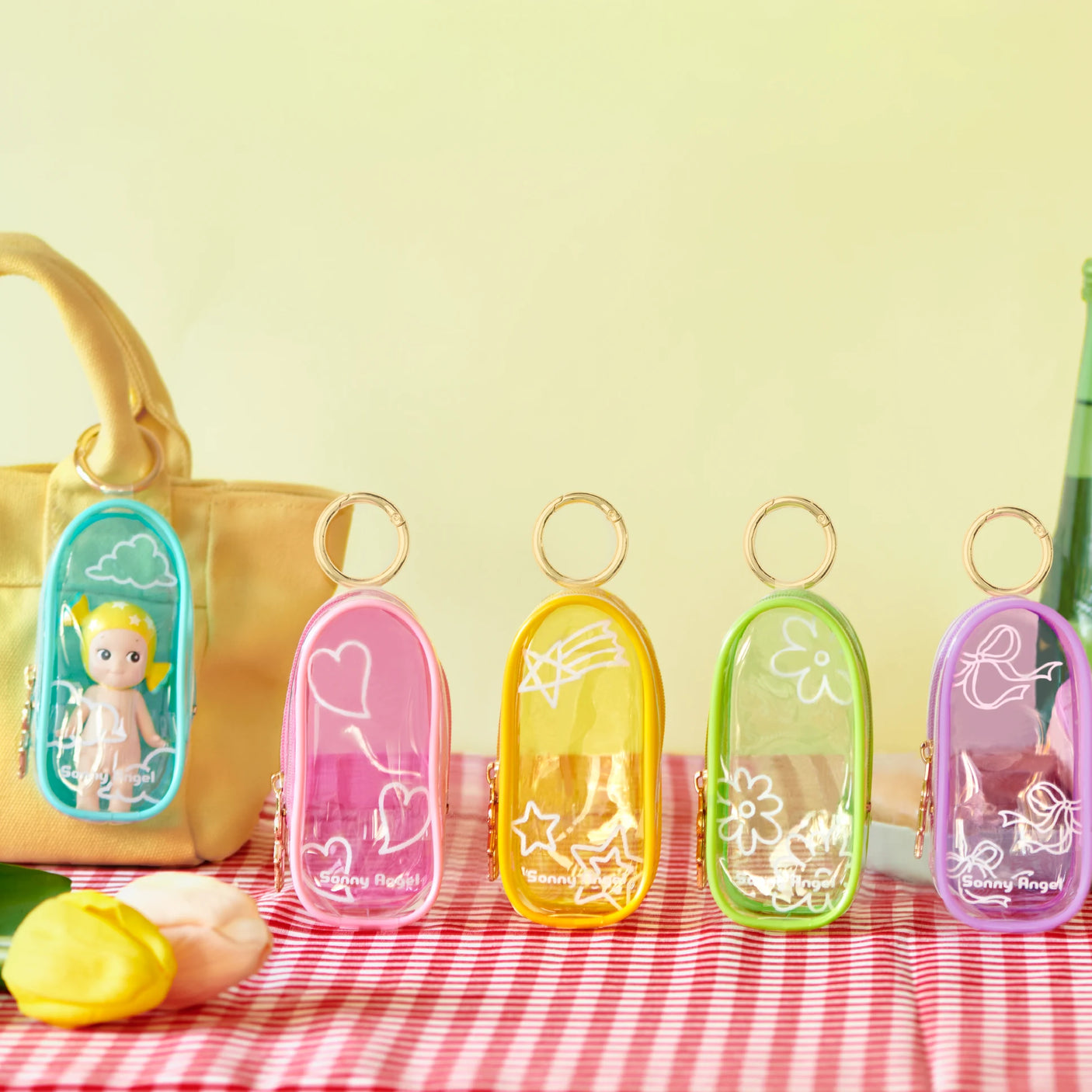 The Sonny Angel My Bestie Case - Purple by Sonny Angel is shown on a red checkered cloth with its white ribbon design, one attached to a beige bag holding a small doll—ideal as a mini figure carry case.