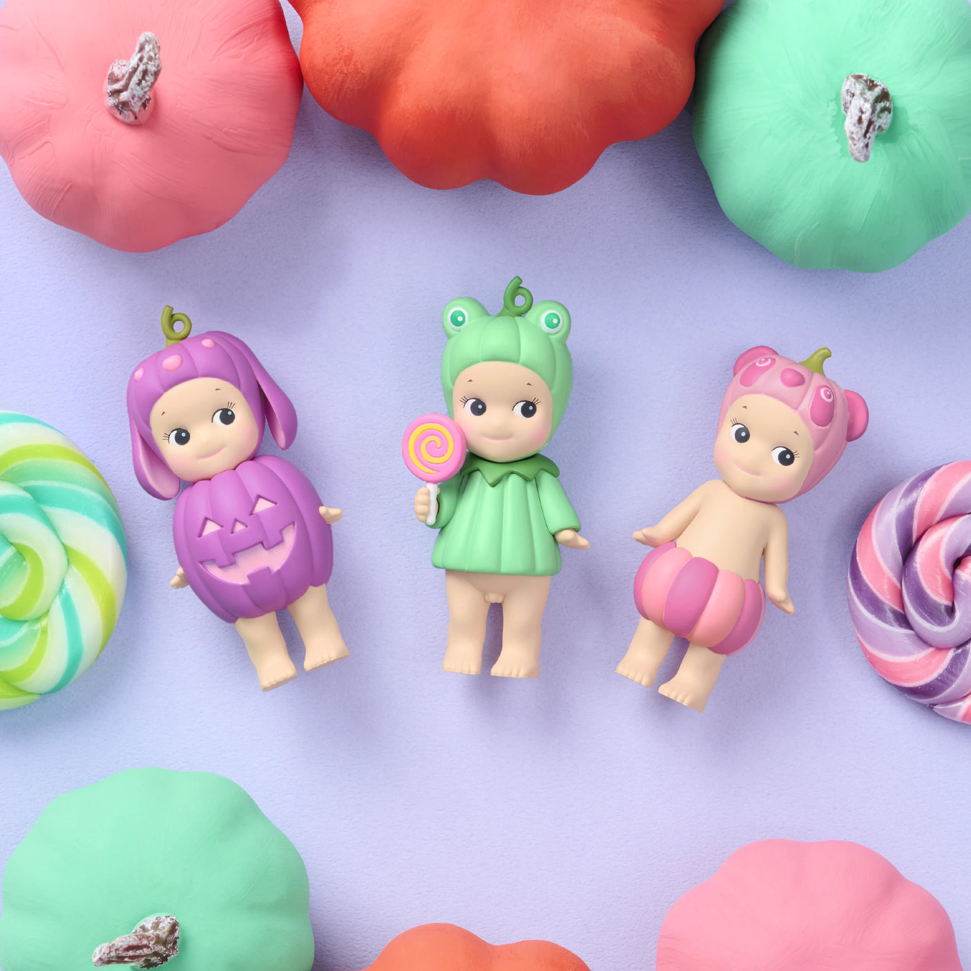 Three Sonny Angel Pumpkin Patch Blind Box dolls by Sonny Angel, dressed in pumpkin, frog, and berry costumes, are displayed on a purple surface with pastel pumpkins and sweets. The Halloween Collection includes a doll holding a lollipop.
