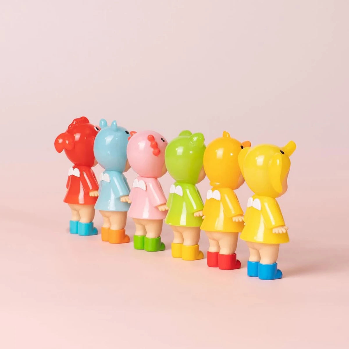 Six Limited Edition Sonny Angel Rainy Day Blind Box figurines in raincoats and boots stand in a row on a pink surface, backs to the camera, capturing the charm of Sonny Angel collectibles.